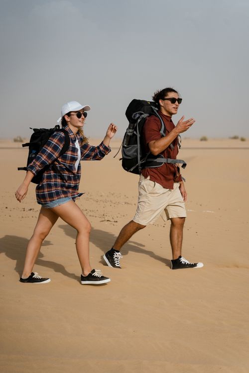 Free Two young adults hiking with backpacks across an open desert on a sunny day, embracing adventure. Stock Photo