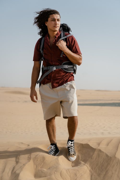 Man in Brown Polo and Carrying his Backpack Standing on Sand 