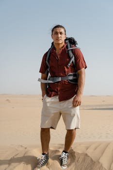 A man stands on sandy desert dunes, wearing a backpack, captured on a clear sunny day.