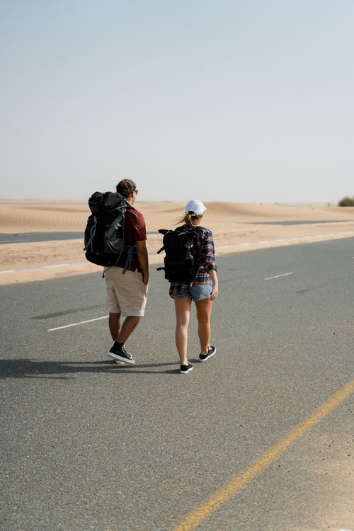 Free A man and woman with backpacks walking on an empty desert road under a clear sky. Stock Photo