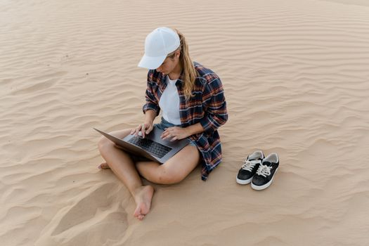 Young woman working with laptop in desert sands, casual attire and remote work concept.