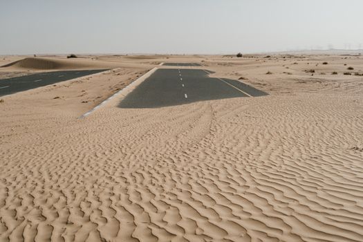 A deserted road partially buried by sand dunes in an arid desert landscape.