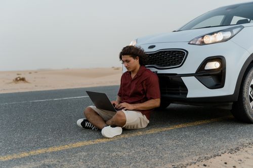 Free A man using a laptop while sitting on the road beside a parked car in a desert setting. Stock Photo