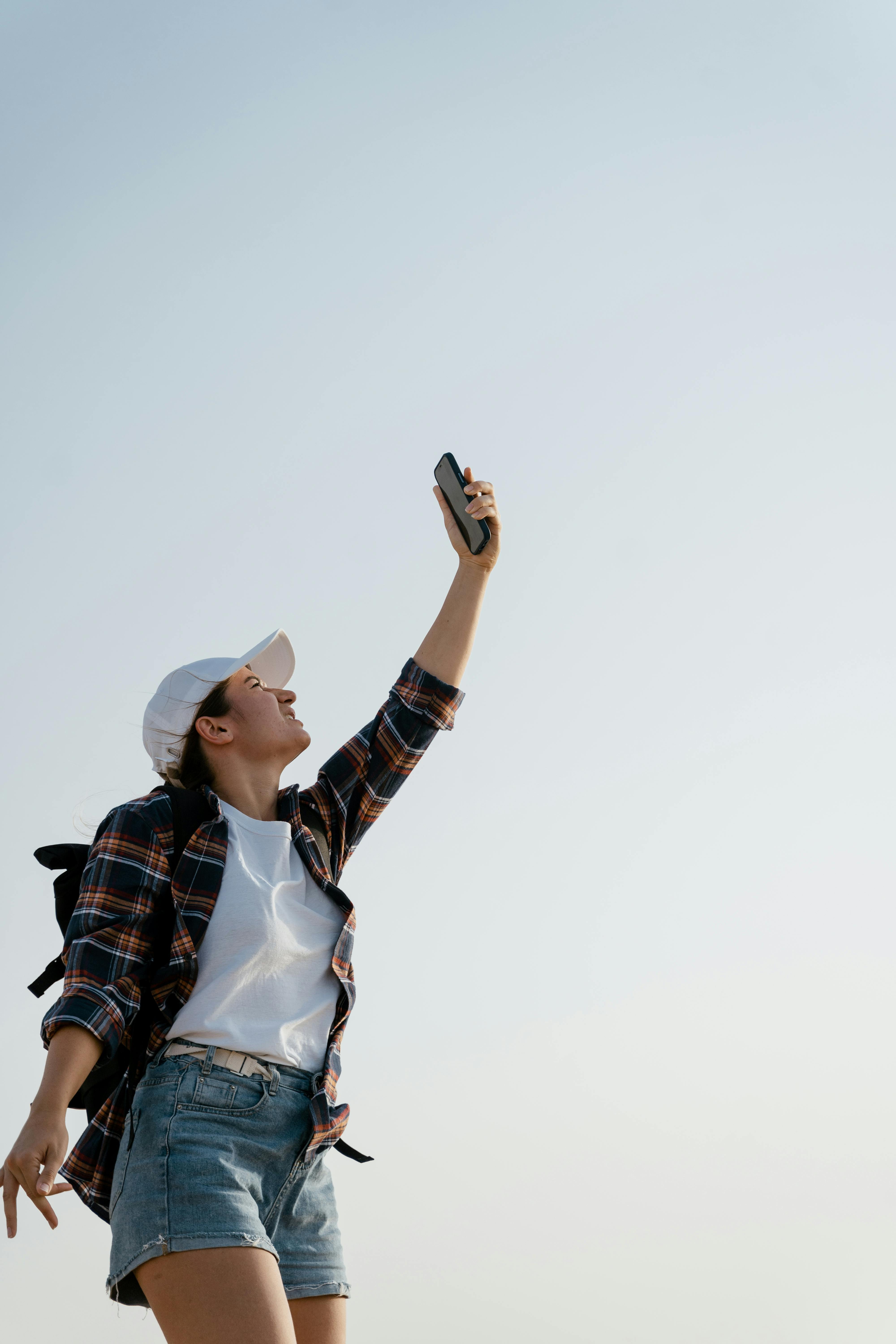 Young woman in casual attire taking a selfie under a clear sky, exuding happiness.