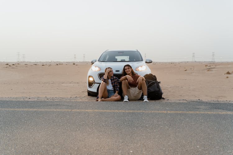 Couple Sitting On The Ground In Front Of Their White Car