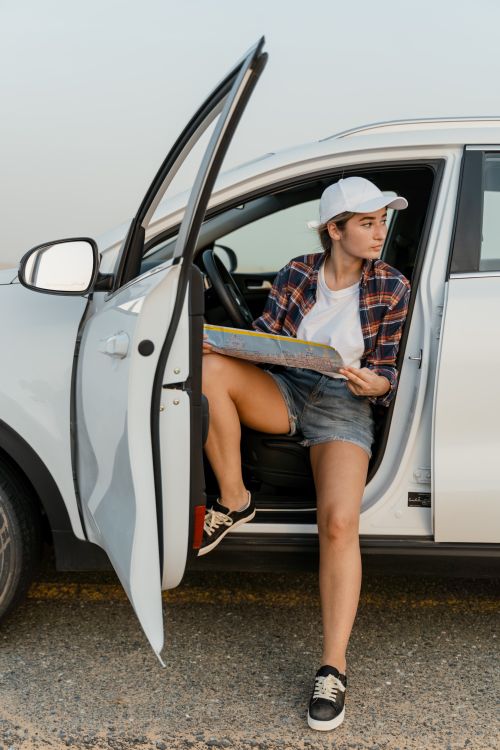 Free Woman in casual attire planning a road trip near a white car, holding a map outdoors. Stock Photo