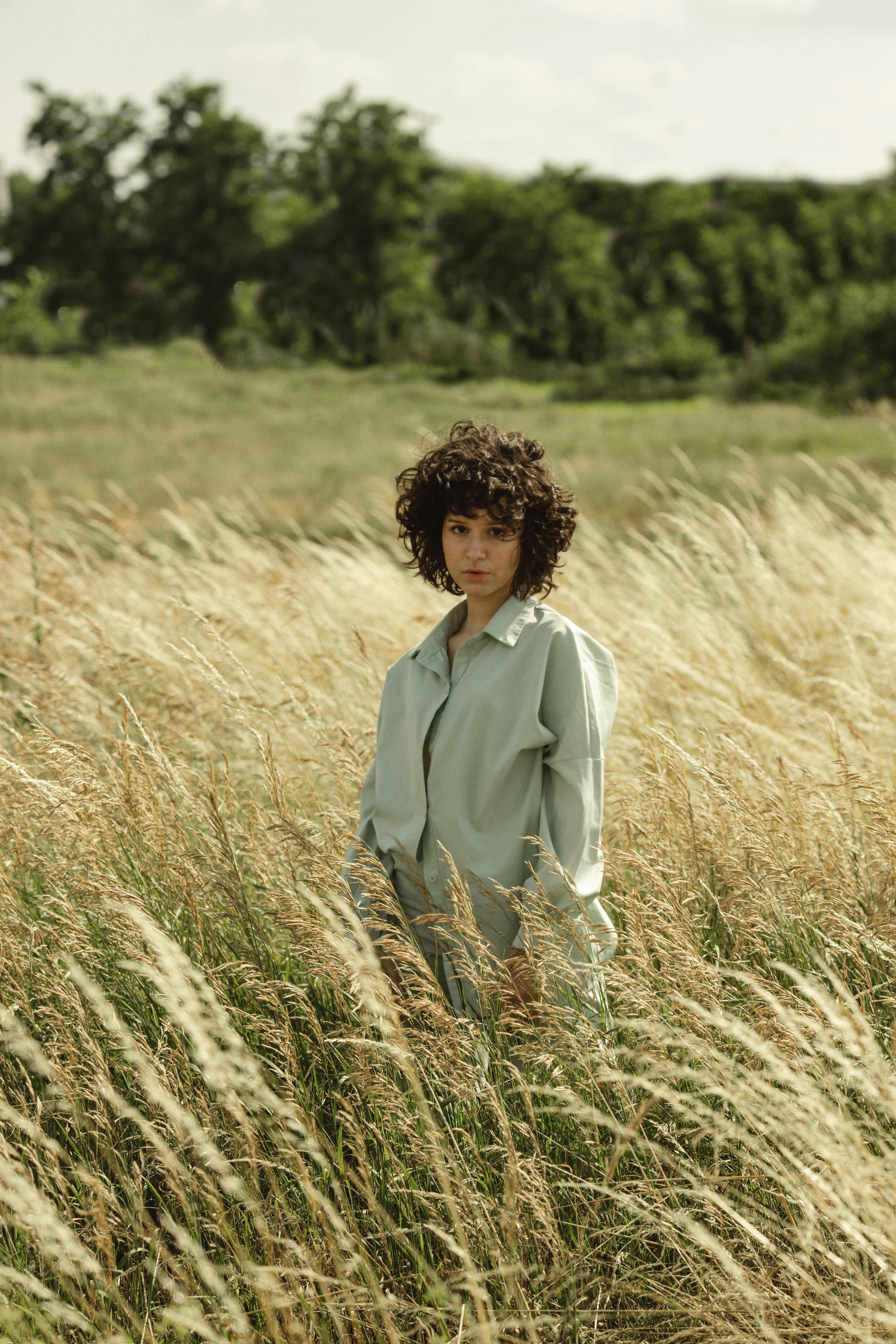 A young woman with curly hair poses gracefully in a verdant field, capturing a serene outdoor scene.