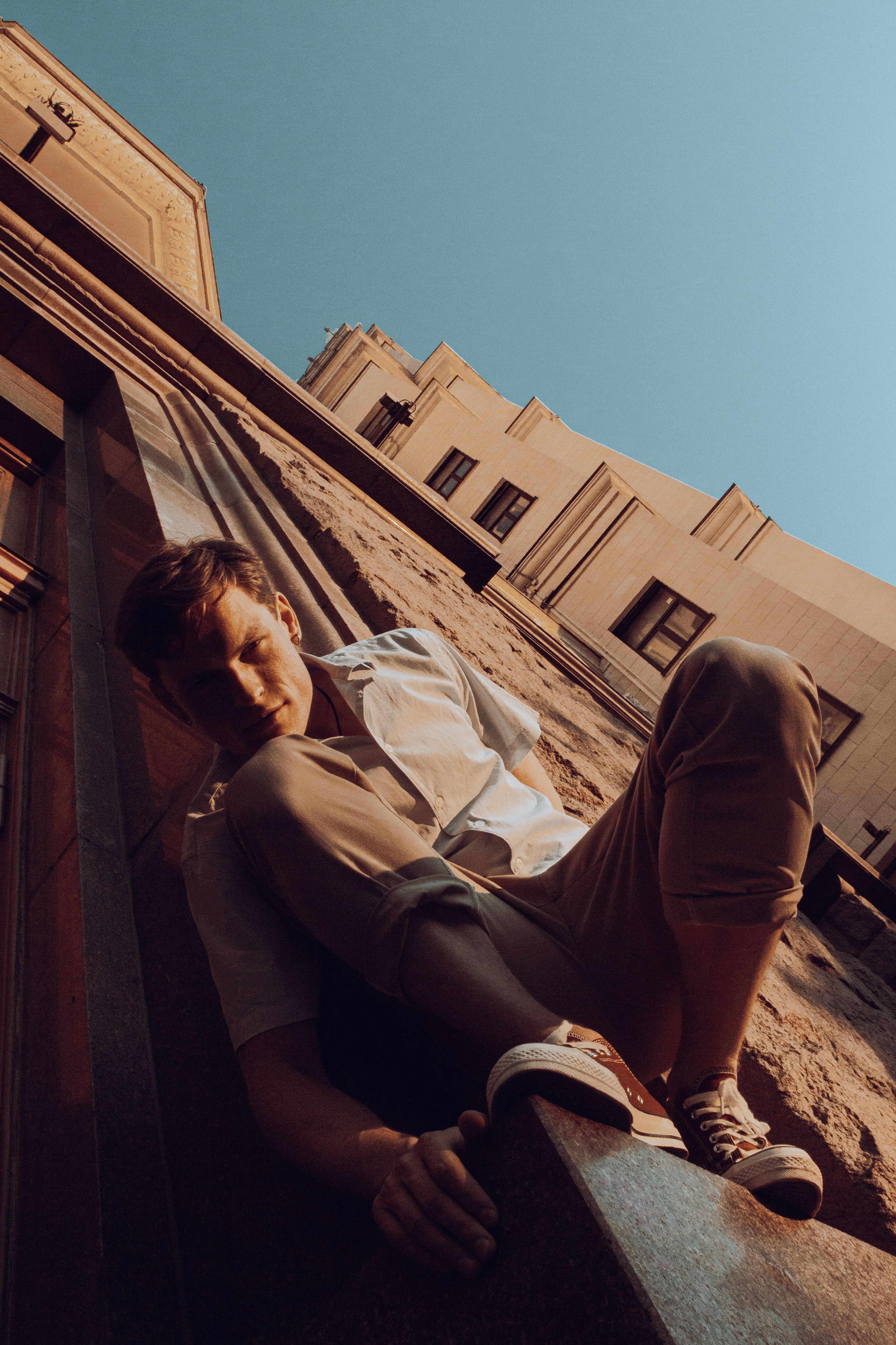 A young man stylishly posing against a building's wall in an urban setting, capturing modern fashion and architecture.