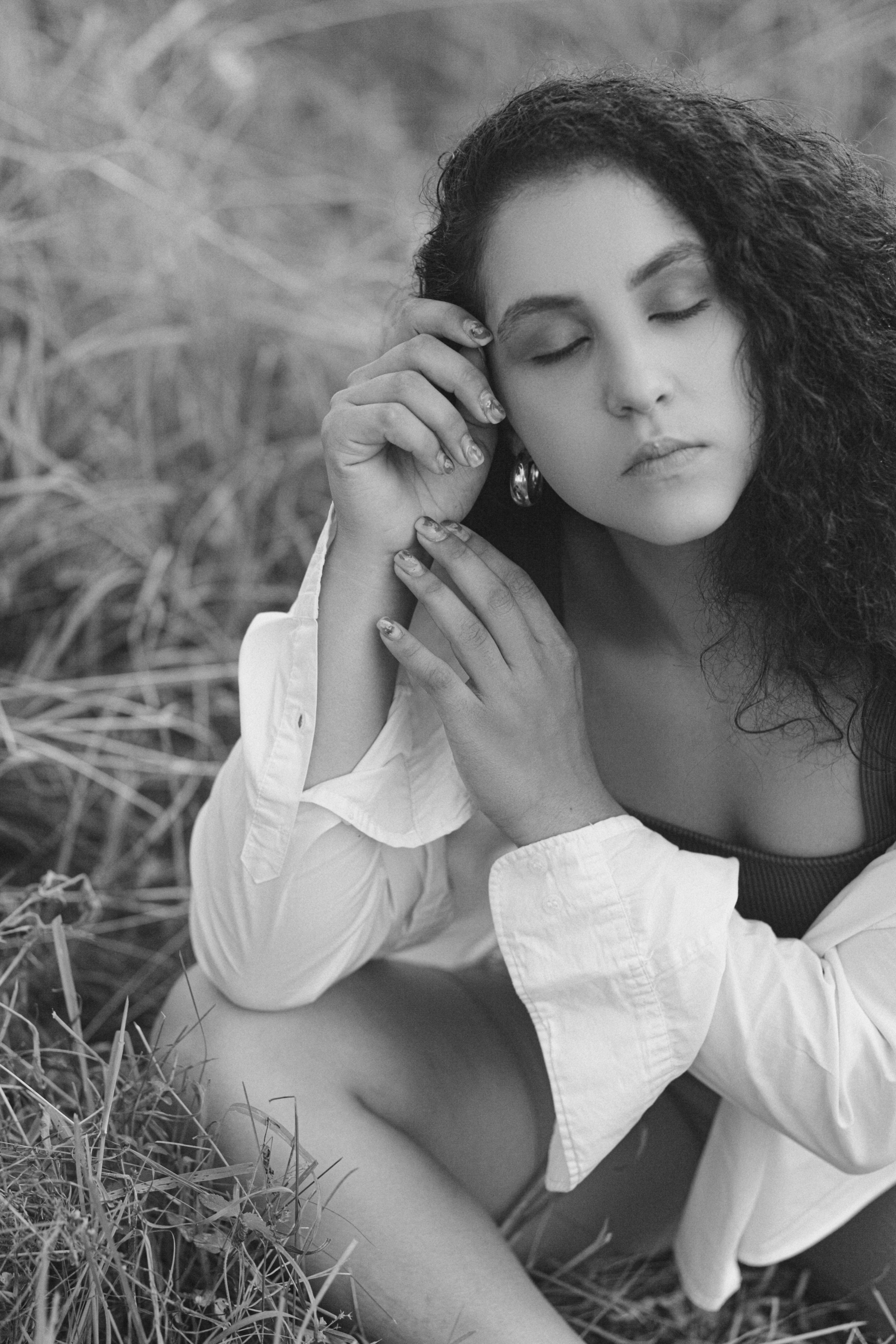 Black and white portrait of a serene woman sitting outdoors, eyes closed, with curly hair and a white shirt.
