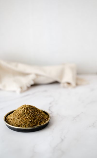 A small glass jar of pale golden-tan beau monde seasoning next to a wooden spoon, onion powder, and celery salt on a kitchen counter.