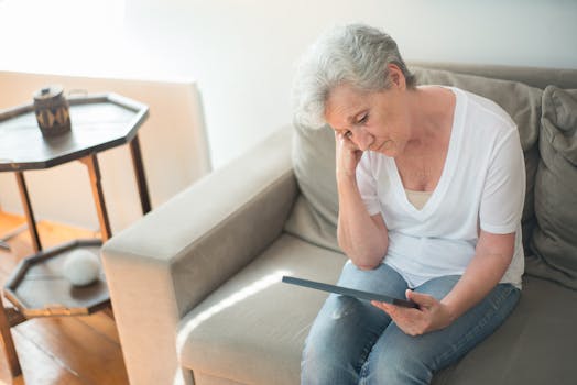 A senior woman sits thoughtfully on a sofa in a bright living room, holding a tablet.