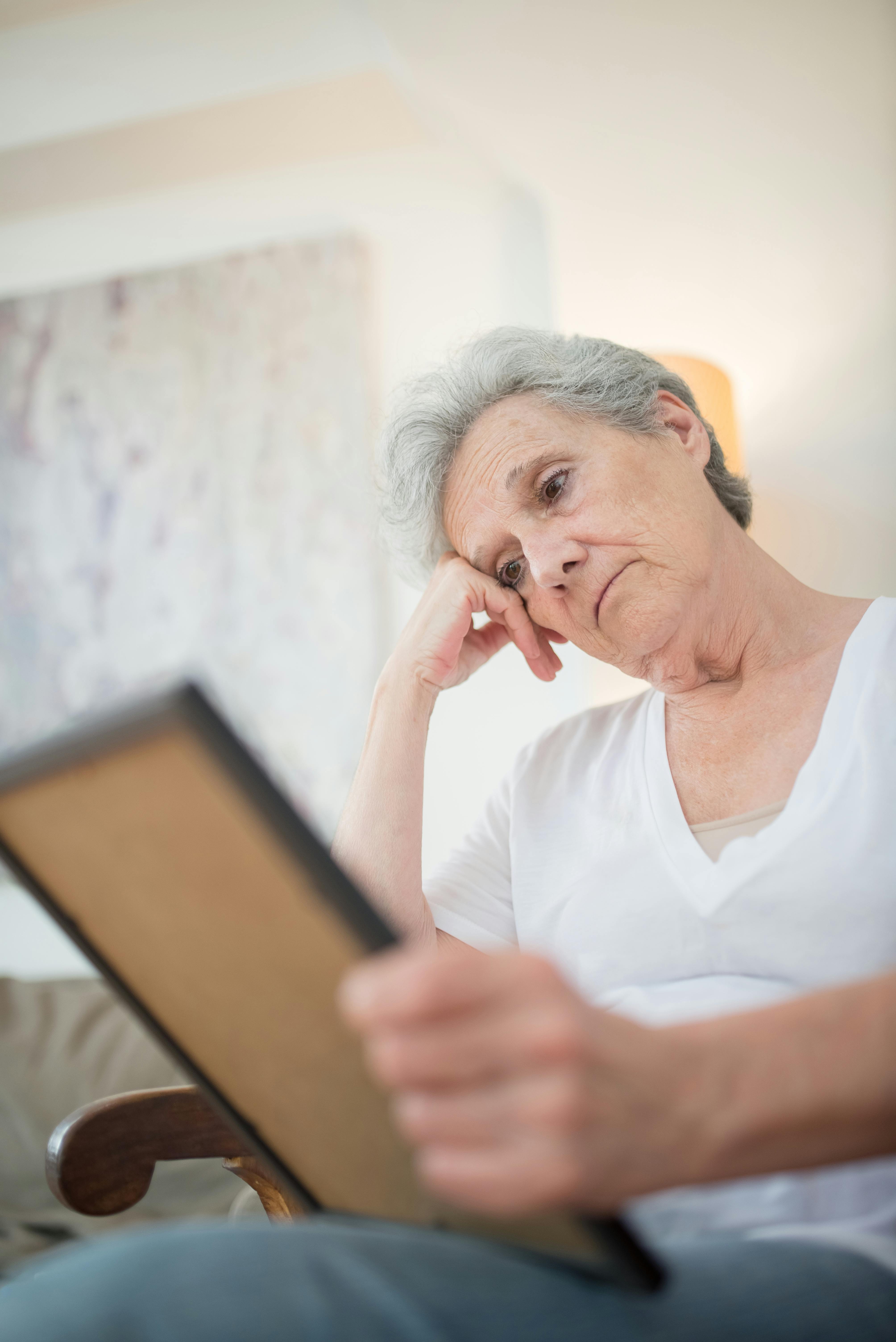 Senior woman in white shirt holding picture frame, reflecting indoors with thoughtful expression.