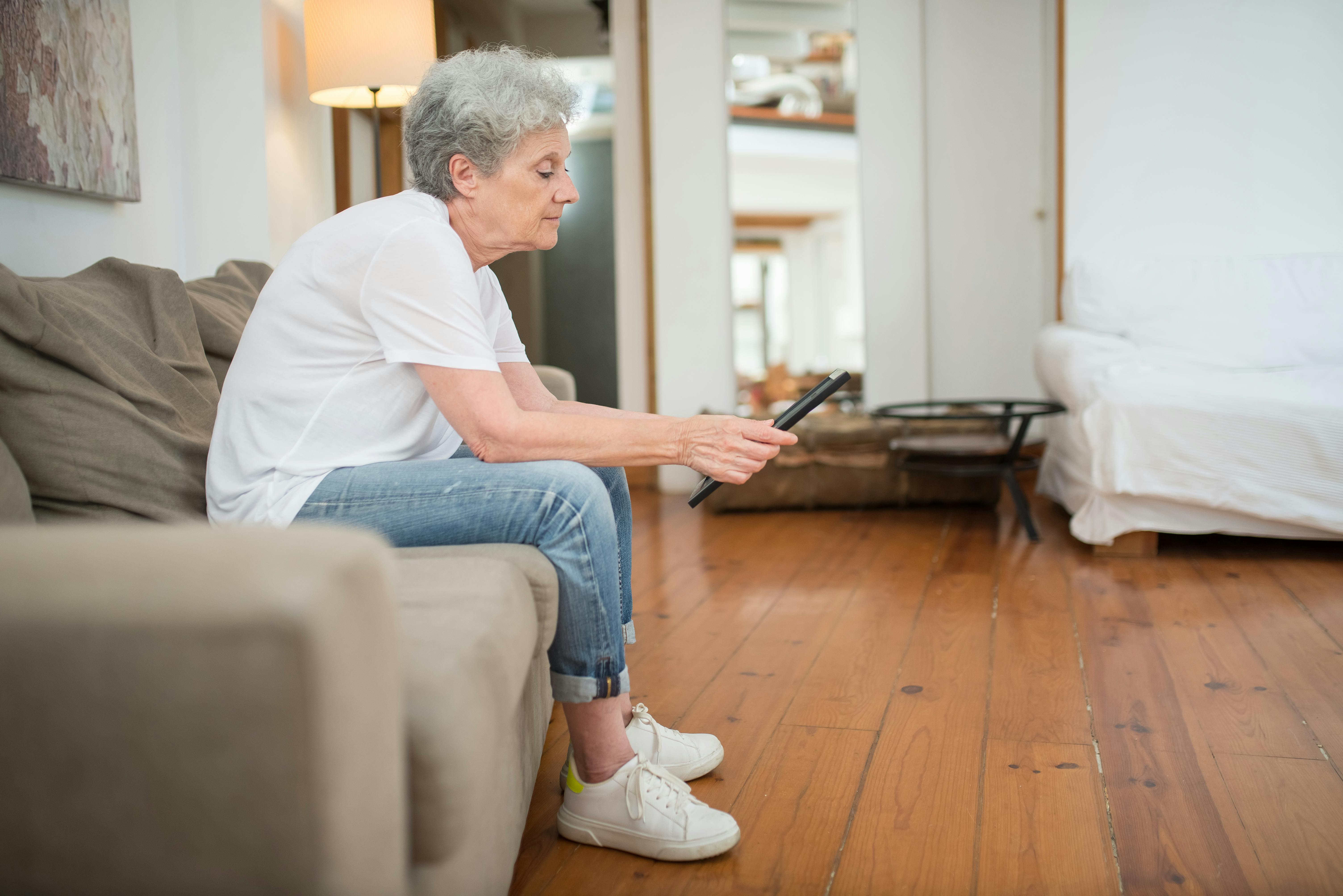Senior woman in casual attire sitting on a couch holding a tablet in a cozy living room.