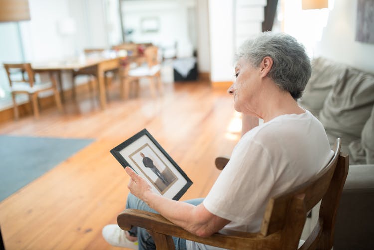 Sad Elderly Woman Sitting On A Chair While Looking At A Picture Frame