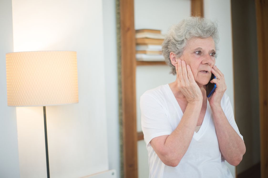 An elderly woman with white hair sits indoors looking worried while speaking on the phone