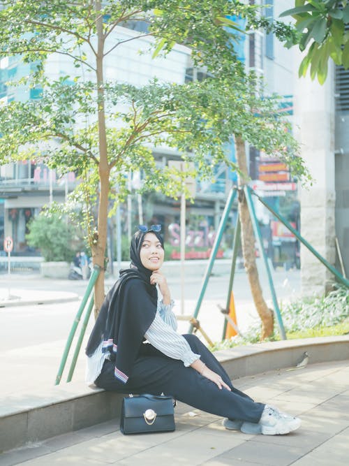 Free A Woman in Black Hijab Sitting on the Side of the Street Near Green Plants while Looking Afar Stock Photo