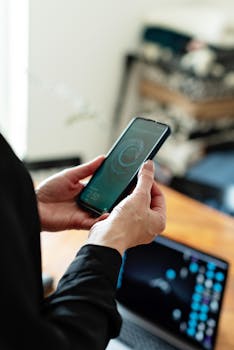 A person holding a smartphone with a visible screen, a laptop in the background on a wooden table.