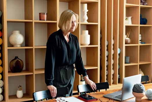 Confident businesswoman standing in a stylish office with wooden shelves, working on a laptop.
