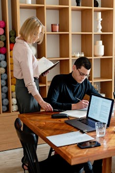 Two colleagues collaborate in a cozy office setting with laptops and documents.