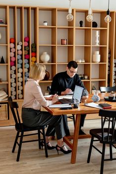Two colleagues working together at a wooden desk in a modern office with yarn display in background.