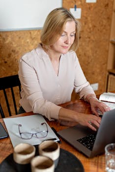 Woman working on laptop at office desk with coffee, glasses, and notebook. Ideal for business and productivity themes.