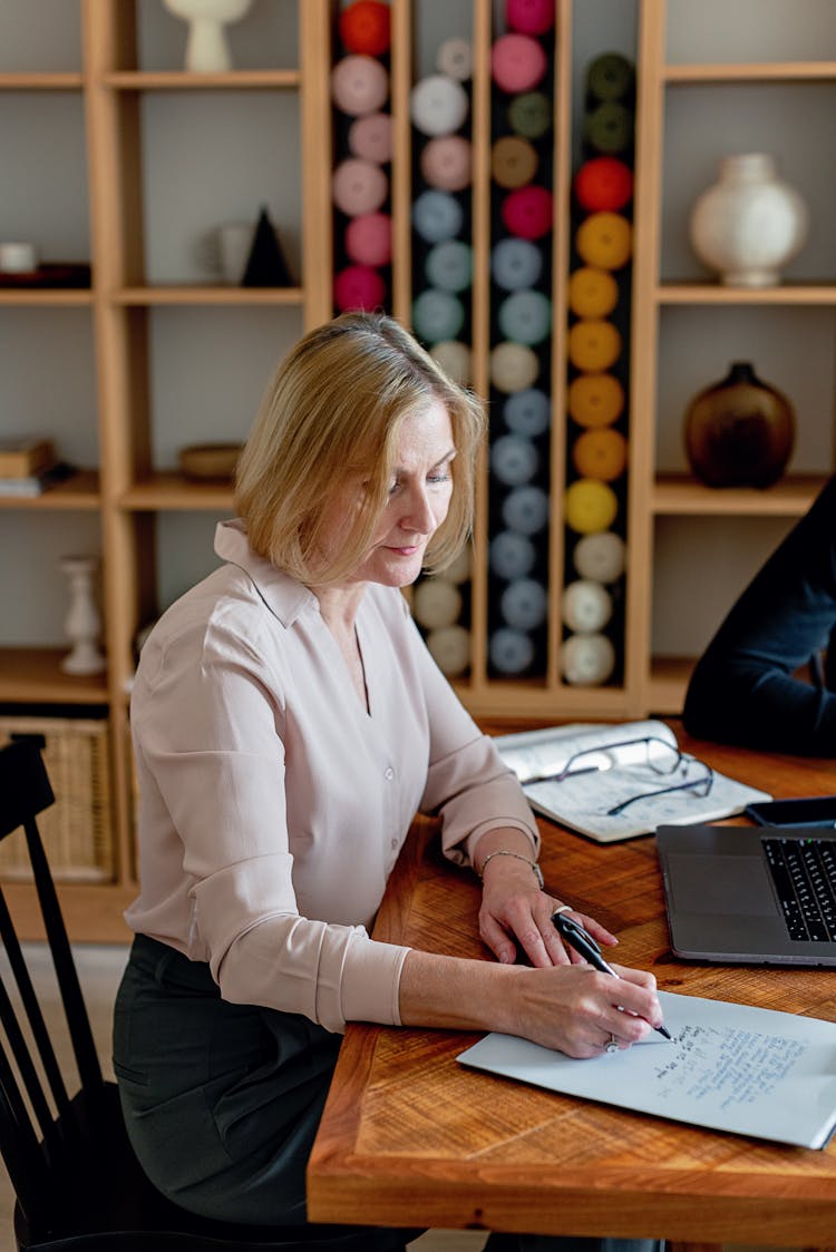 A Mature Woman Sitting At A Wooden Desk Writing On Paper