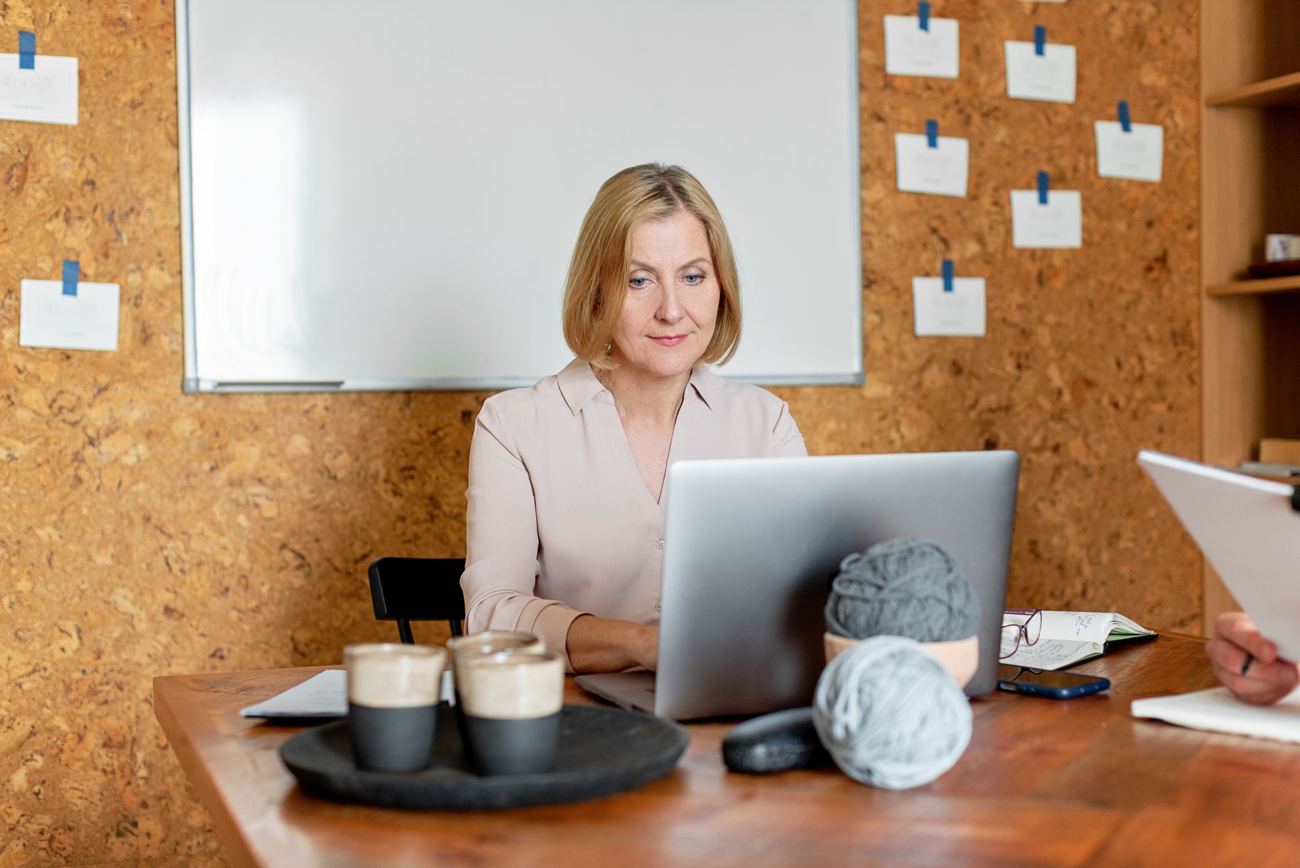 A blonde woman working on her laptop in a cozy office setting with yarn balls and coffee.