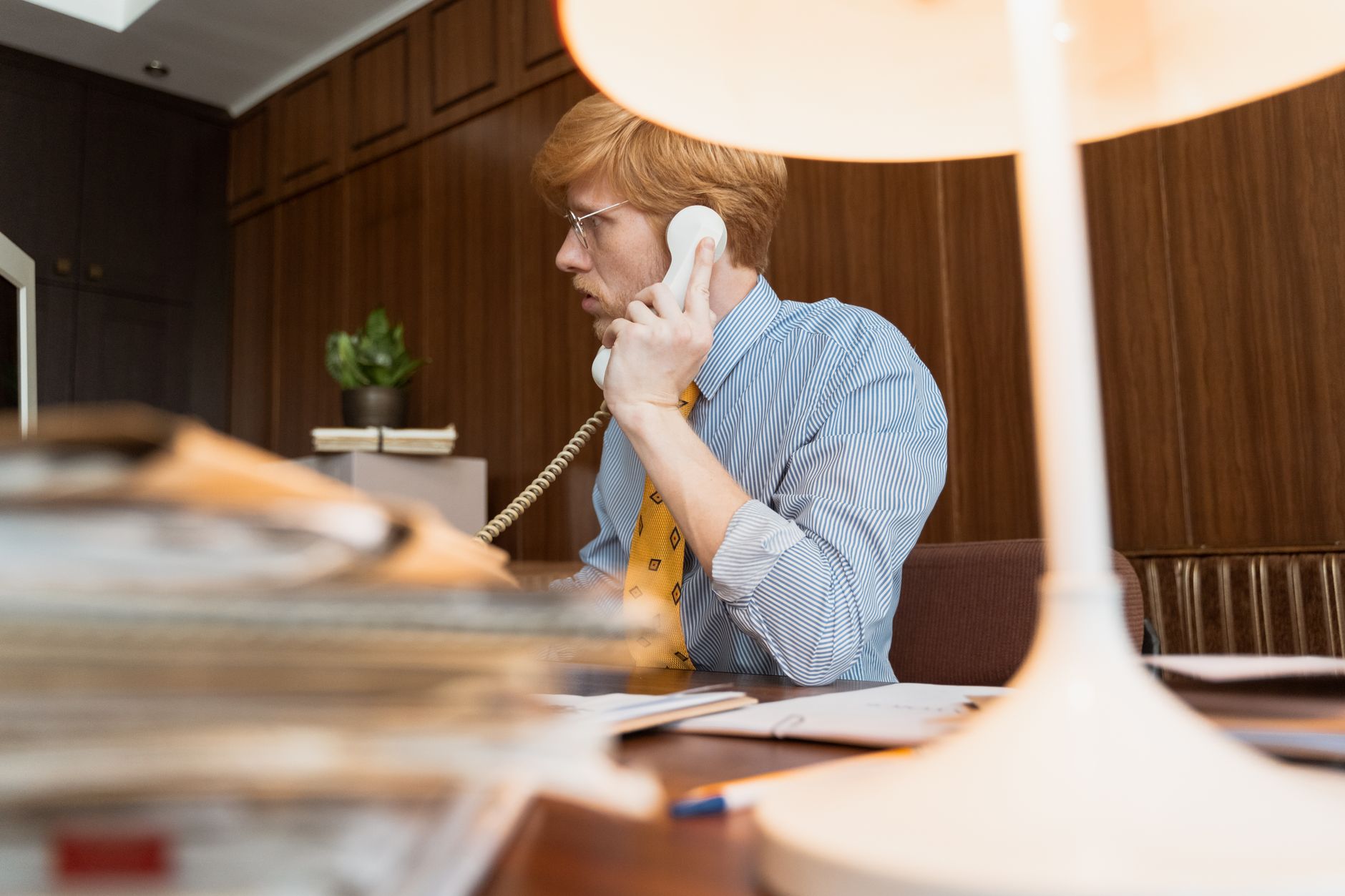 Professional man with eyeglasses talking on a vintage telephone in an office setting.