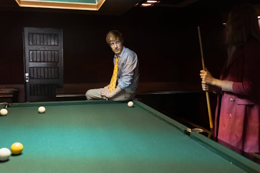 A man in a yellow tie sits on a billiard table while a woman holds a cue stick in a dimly lit room.