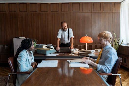 Three colleagues having a discussion in a vintage-style office with a typewriter and wooden paneling.