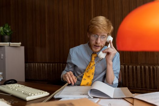 Caucasian man with red hair in retro office, talking on vintage telephone.