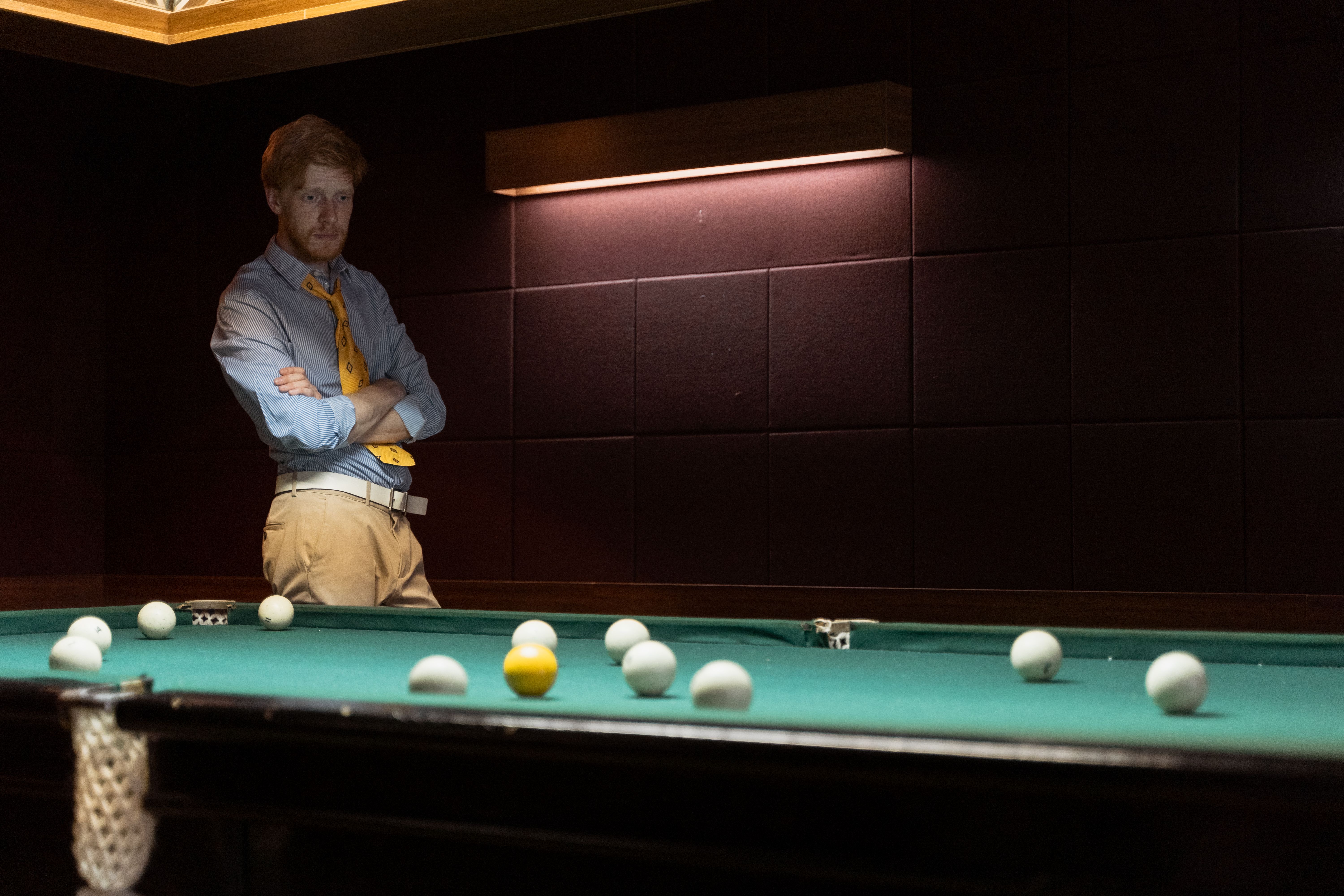 A man with a yellow tie and arms crossed stands by a billiards table, ready to play.