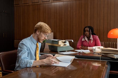 Man in Blue Dress Up Shirt Browsing Papers Woman in Pink Coat Looking at Man 