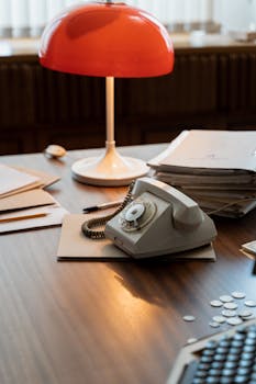 Retro desk setup with rotary phone, red lamp, and documents suggesting a classic office vibe.