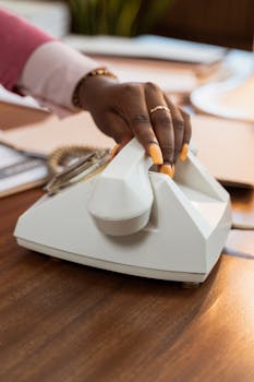 Close-up of a hand with nail art picking up a vintage telephone on a desk.