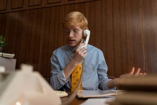 Man with blond hair in business attire making a phone call in a retro-styled office setting.