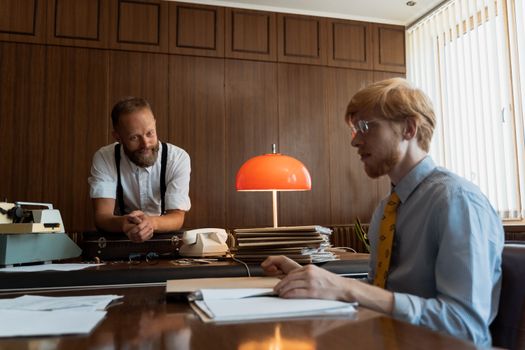 Retro office scene with two men discussing work at a desk, featuring a typewriter and documents.