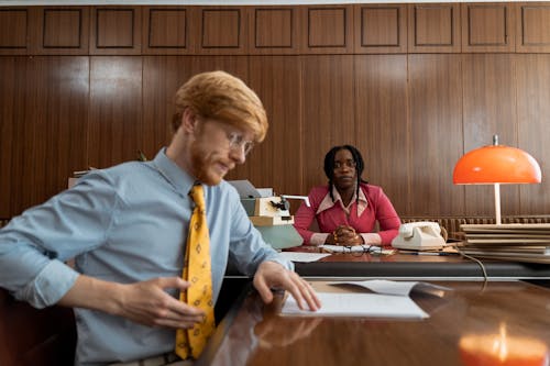 Man in Blue Dress Up Shirt Browsing Papers Woman in Pink Coat Looking at Man 