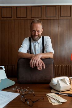 Elderly man with suspenders smiling indoors, leaning over a desk.