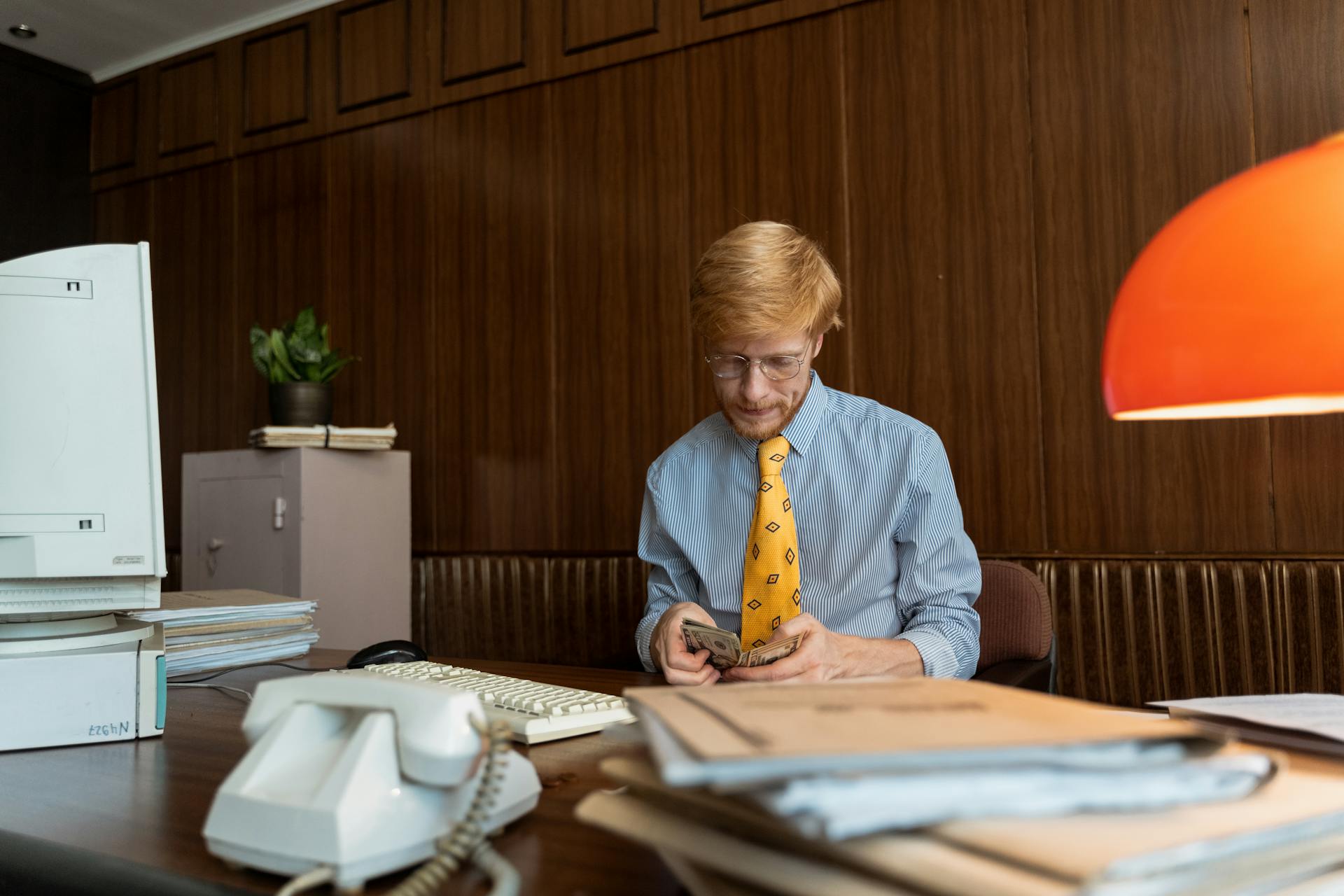 A professional counting cash at a retro office desk featuring a computer and files.