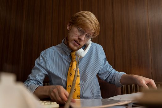 Red-haired businessman in glasses multitasking at his desk using a telephone and working on documents.