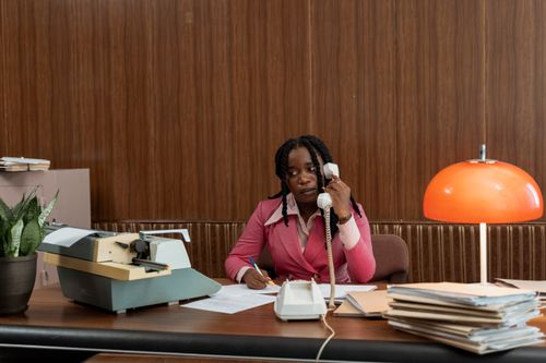 Free Businesswoman in retro office attire on phone call with typewriter, papers on desk. Stock Photo