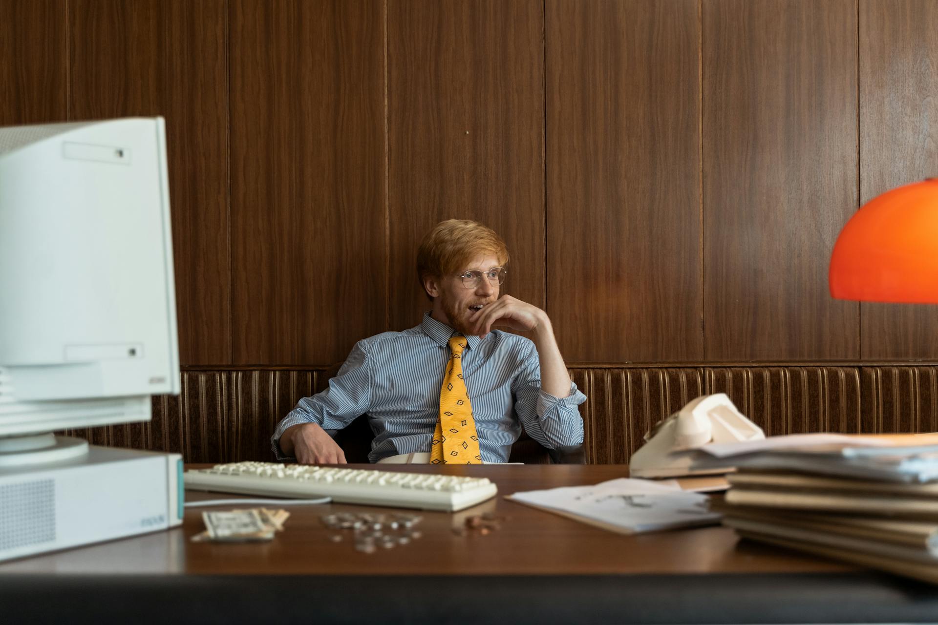 A man in a vintage office setting, looking contemplative with old computer and files.