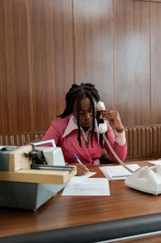 Businesswoman in vintage attire sitting at a desk, taking notes and on a phone call.