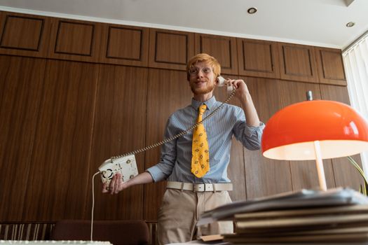 Caucasian man with red hair talking on a vintage phone in a classic office setting.
