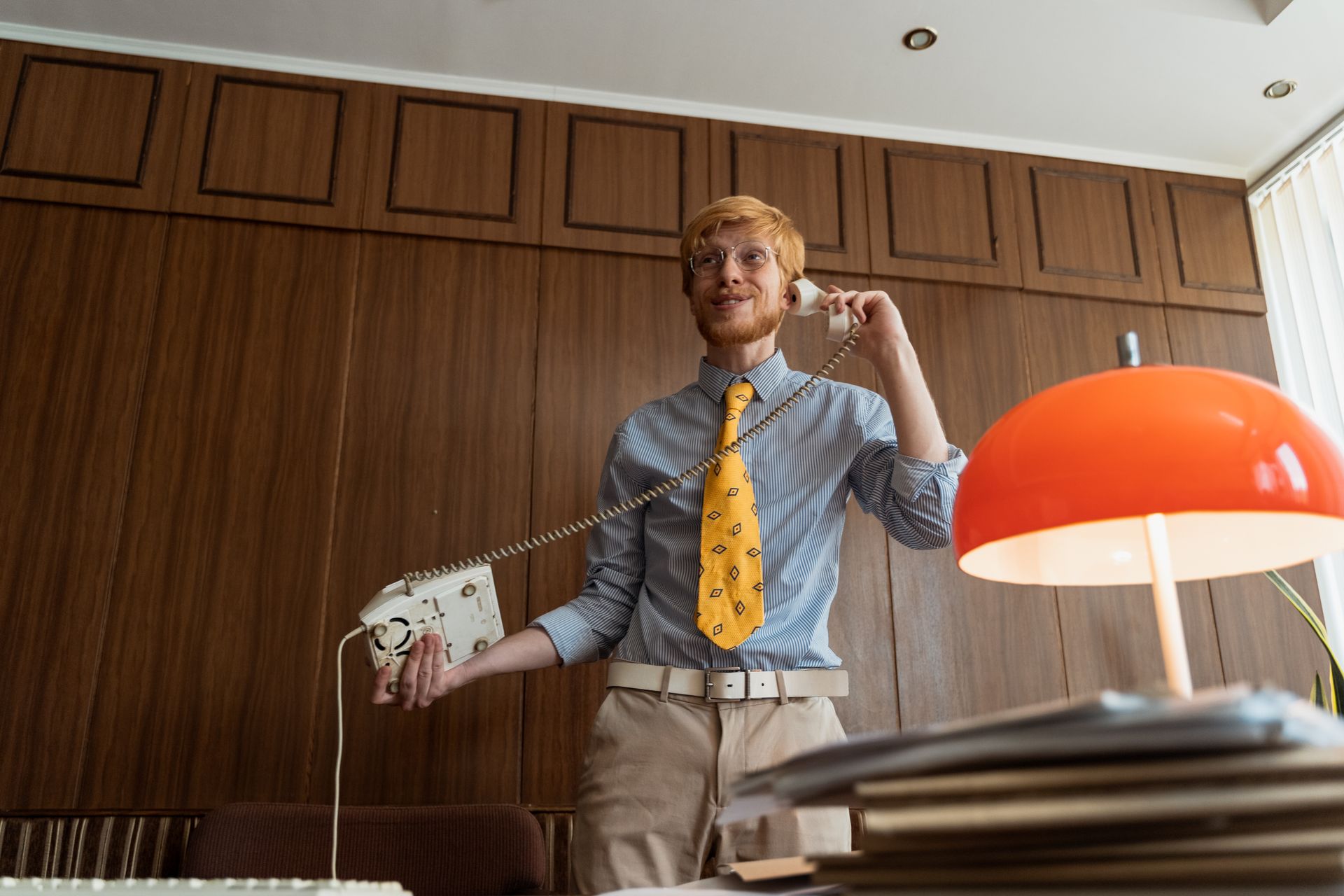 Caucasian man with red hair talking on a vintage phone in a classic office setting.