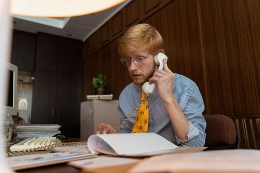 A professional man in a vintage office setting talking on a rotary phone.