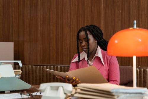 Free A woman in a vintage office setting reading a folder, surrounded by retro items. Stock Photo