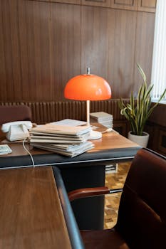 A vintage office setup featuring a stack of documents, orange lamp, and classic wooden furniture.