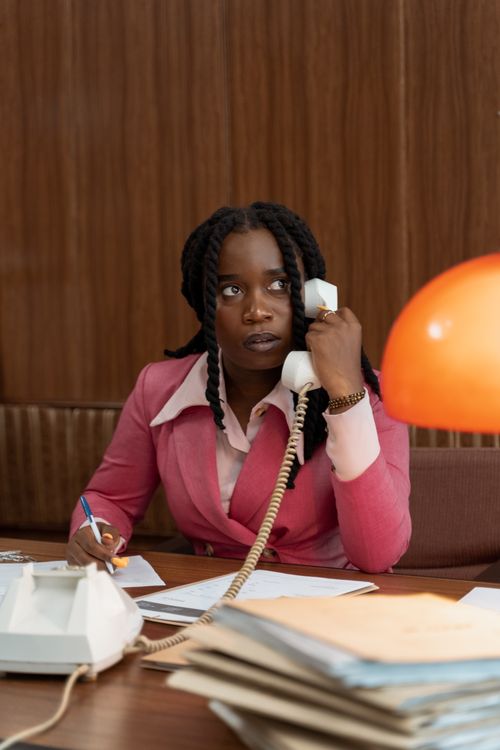 Free African American woman in a pink suit talking on a vintage telephone at an office desk. Stock Photo
